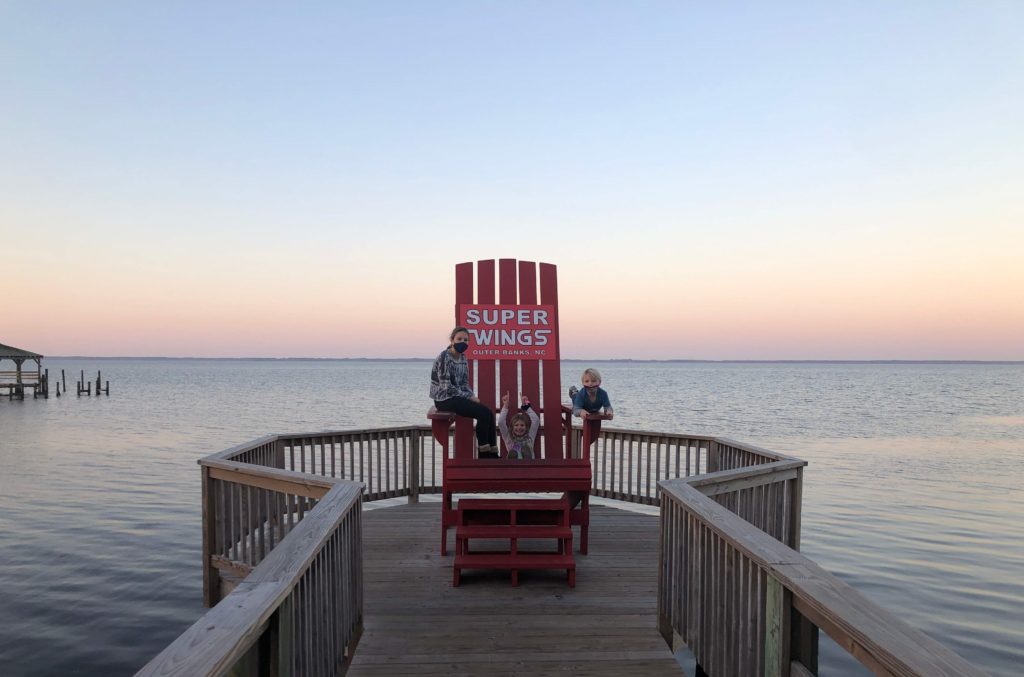 three smiling children pose in a giant Adirondack chair on the boardwalk in Duck, North Carolina