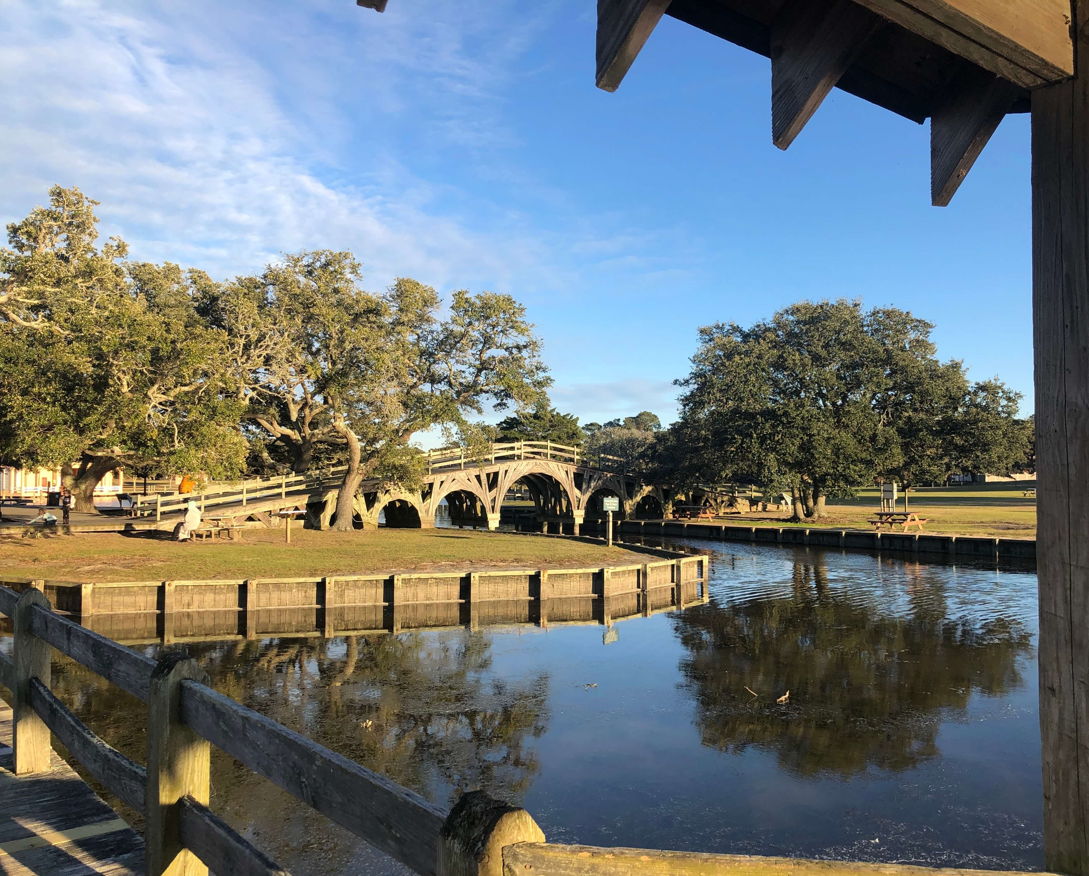 a wooden bridge lined with trees  crosses an inlet while picnic tables surround