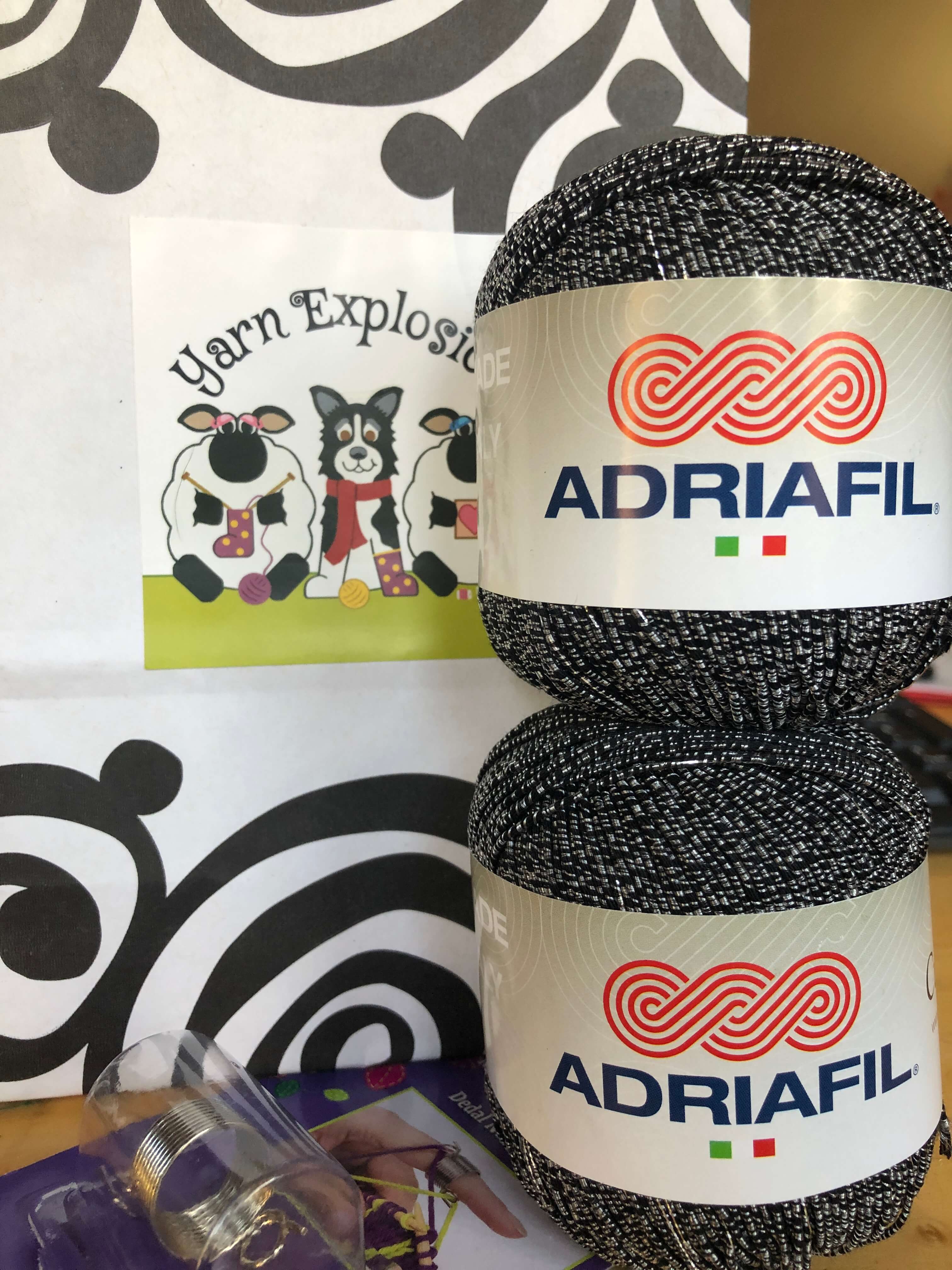 a close shot of a bag reading "Yarn Explosion" next to a textured ball of reflective yarn (Adriafil Cupido)