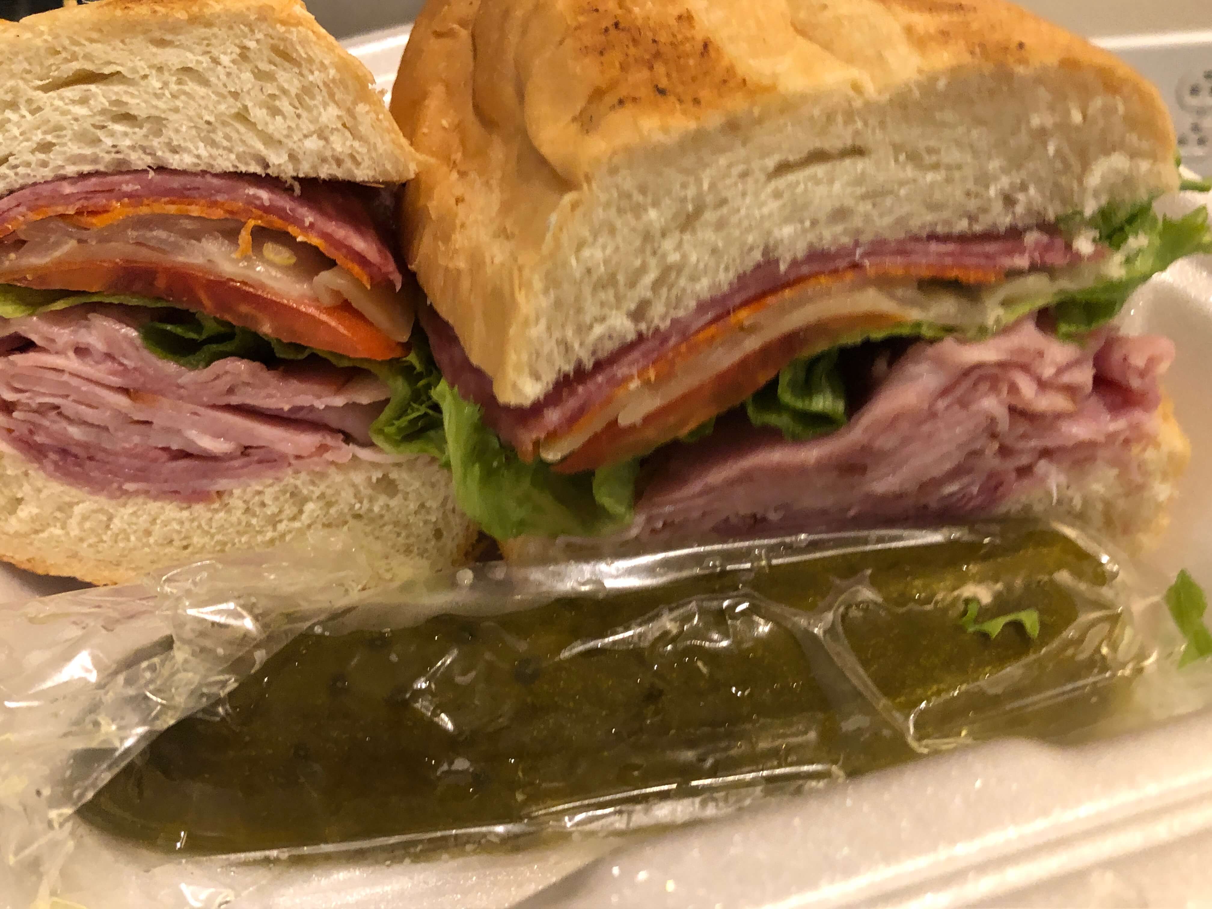 a sandwich with various Italian meats, lettuce, and tomato on French bread with a pickle