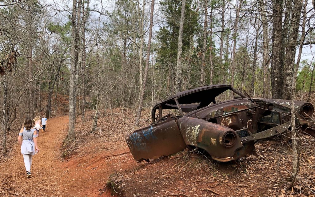 three children hike a trail past a rusting, broken antique car in the forest