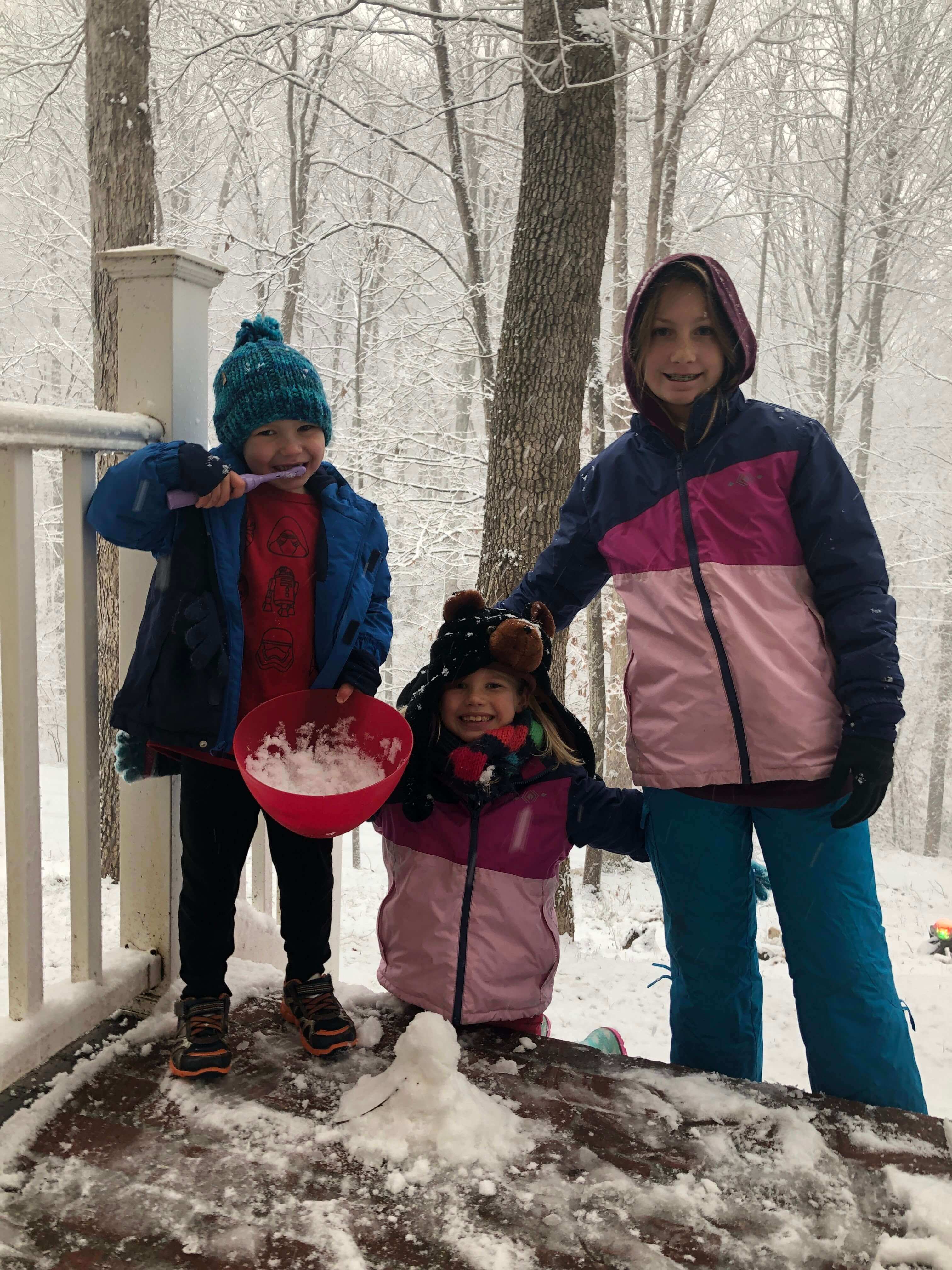 three children in winter gear pose with a tiny snowman and bowl of snow ice cream