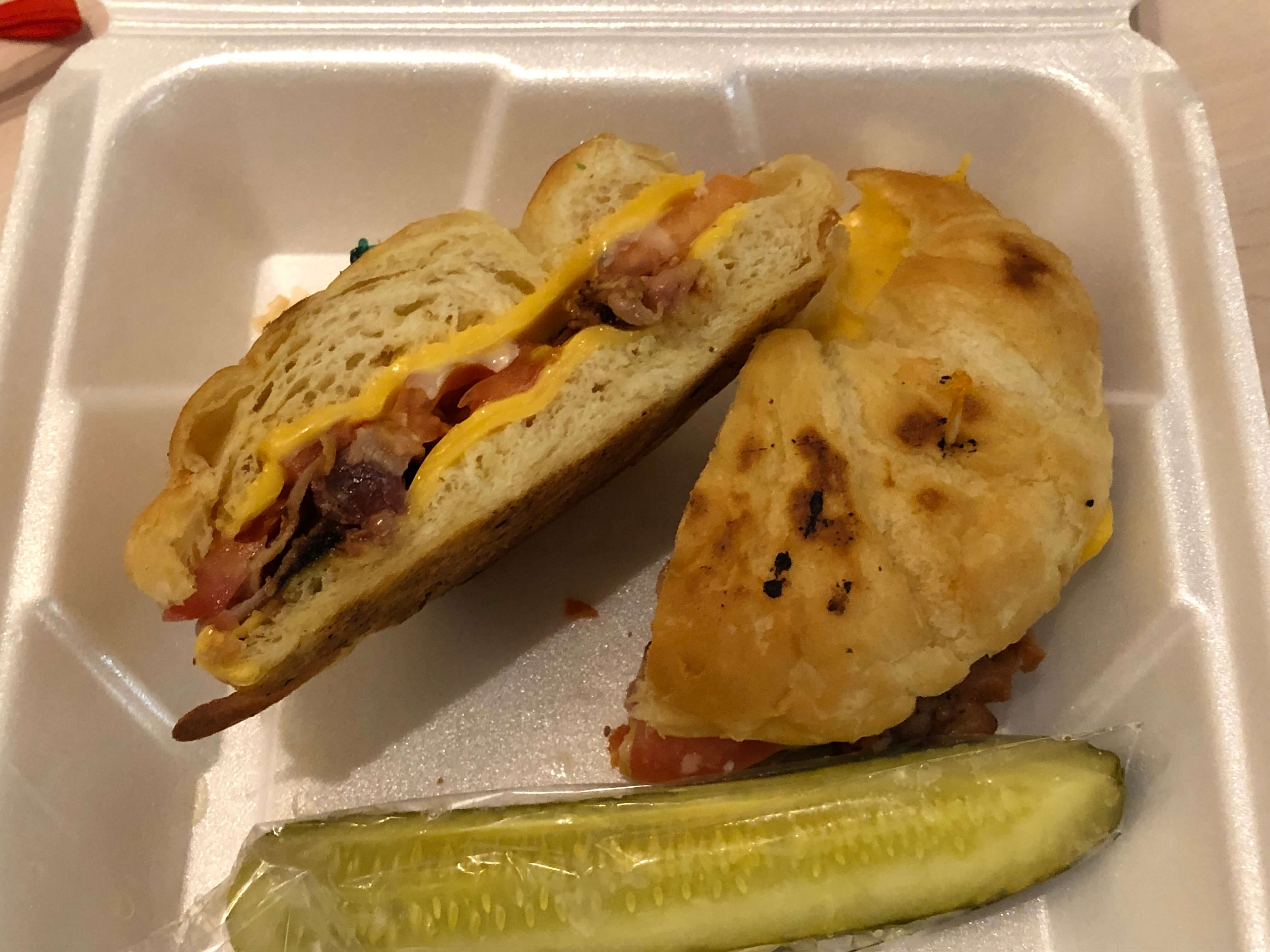 a croissant sandwich with tomato, cheese, and bacon with a pickle in Styrofoam clamshell