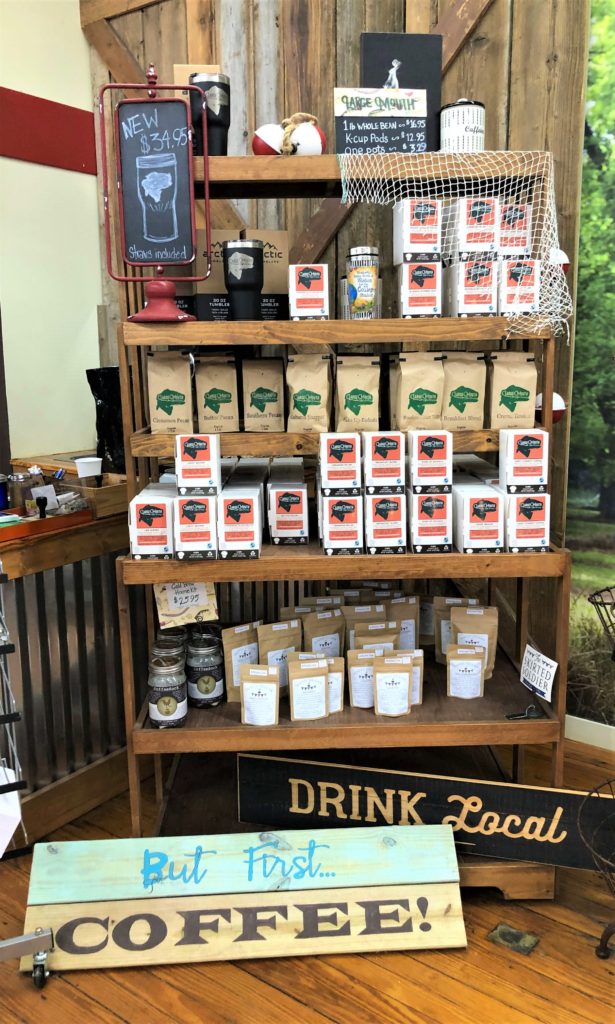several varieties of bagged coffee are stacked above signs reading "Drink Local" and "But First... Coffee!"