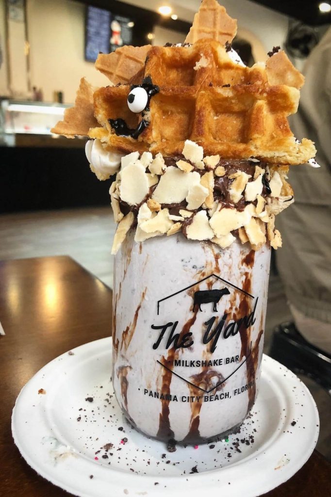 a smiling dinosaur-shaped waffle with sugar cone spikes smiles while sitting on top of a glass jar full of milkshake