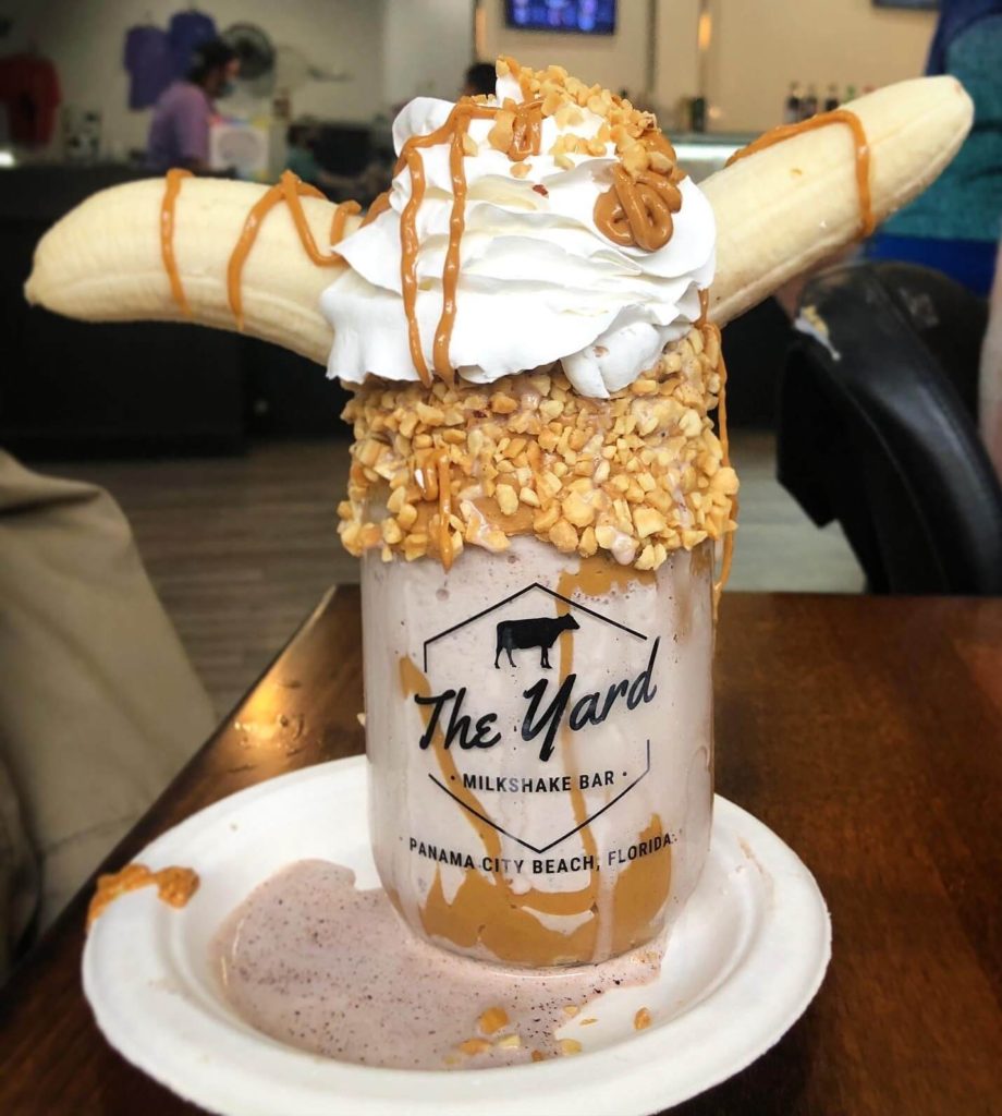 Two banana halves poke out either end of a milkshake jar covered in melted peanut butter, chopped peanuts, and whipped topping.