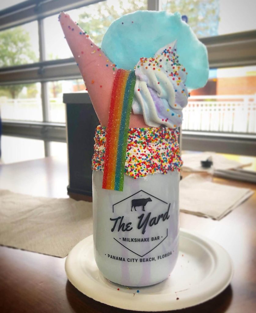 Rainbow sprinkles top a pink chocolate-dipped sugar cone, cotton candy cloud, rainbow chewy candy, and multi-color whipped cream dollop. All are perched atop a glass jar full of milkshake.