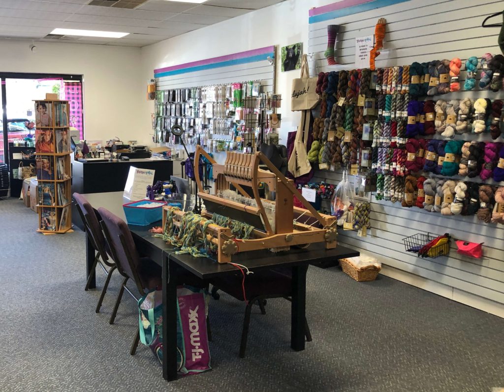 a loom with a work in progress sits in the center of a yarn shop, surrounded by yarn