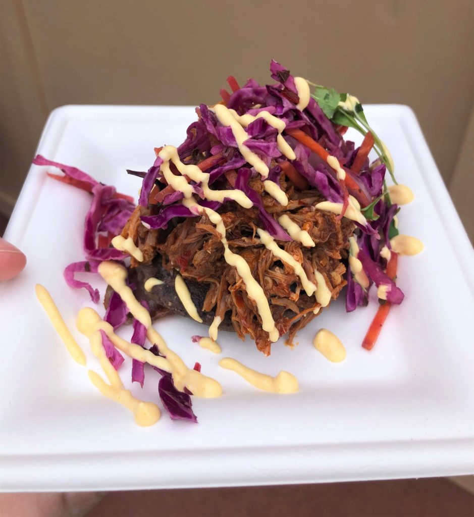 a blue corn pupusa stuffed with cheese and topped with shredded pork, chile sauce, red cabbage slaw, and drizzled with crema