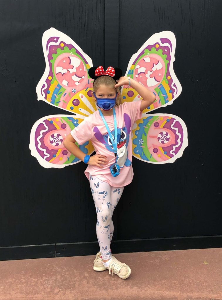 a smiling girl in Mickey Mouse ears poses in front of a wall painted with butterfly wings