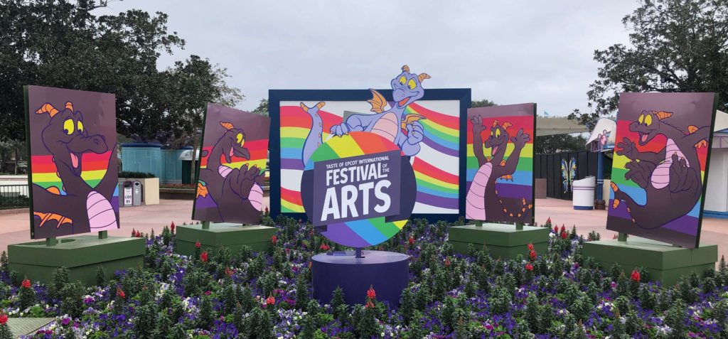 Five paintings of a playful dragon and rainbow line a garden. The center one reads "Festival of the Arts."