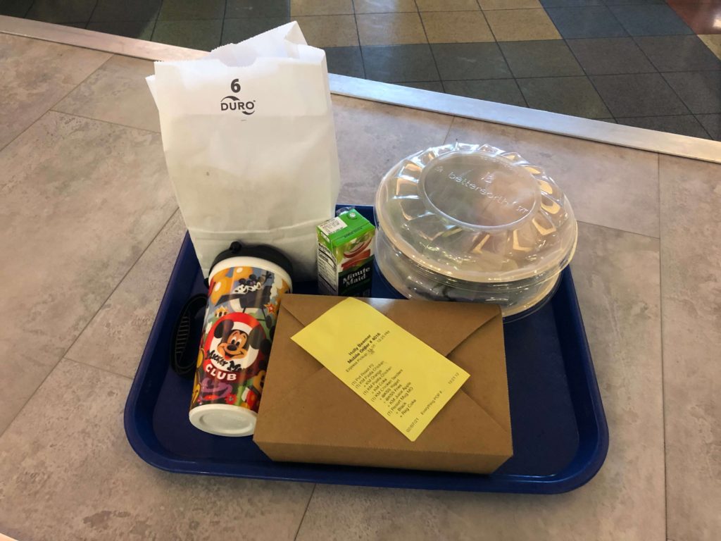 a meal tray with three meals, a paper bag, a reusable mug, and an apple juice box