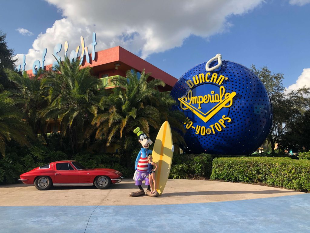 Goofy, wearing a swimsuit and trunks and holding a surf board, stands in front of a two-seater car and giant yo-yo at Pop Century Resort