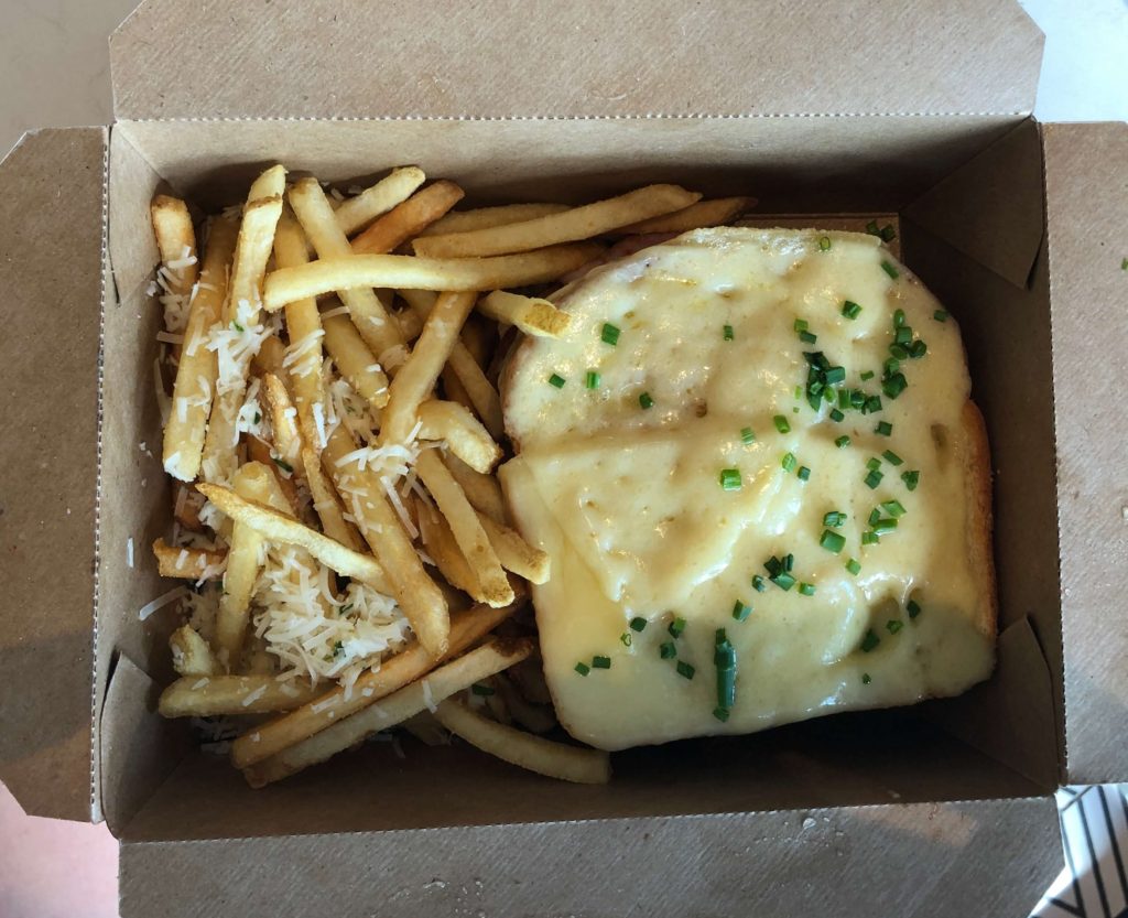 a to-go meal container holds a croque monsieur sandwich covered in Swiss cheese and green onions with French fries covered in Romano cheese and herbs