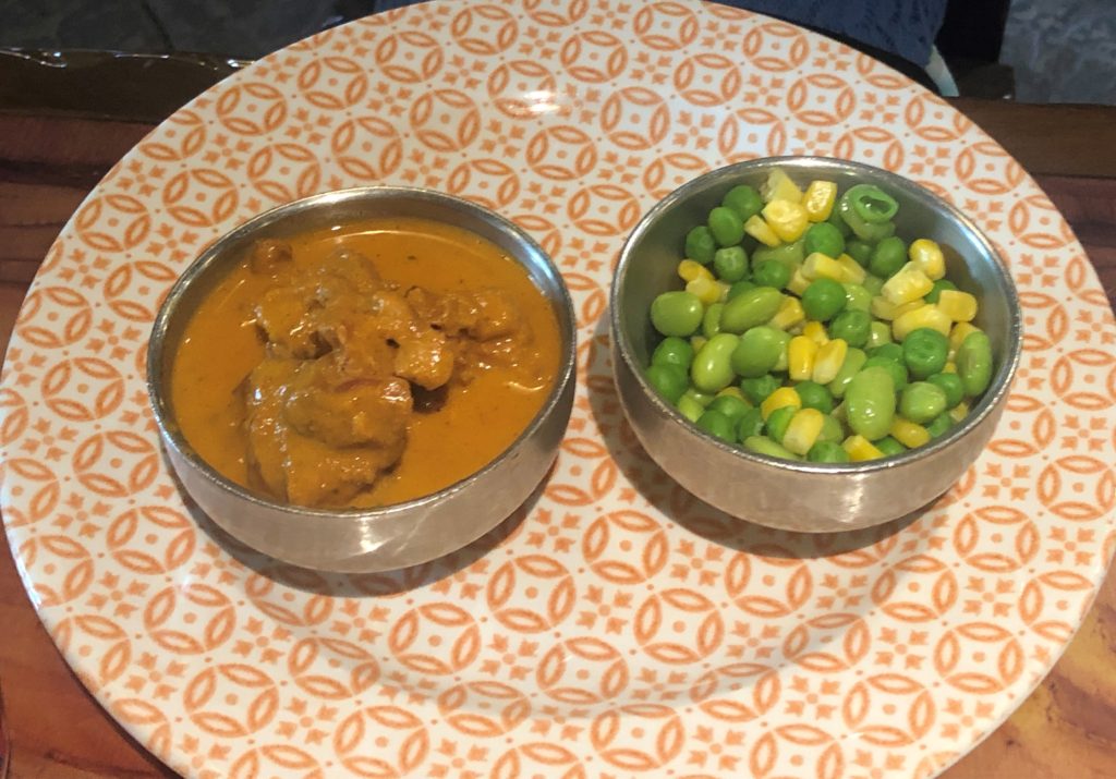 a metal bowl of butter chicken and a metal bowl of mixed peas, corn, and lima beans on a patterned plate