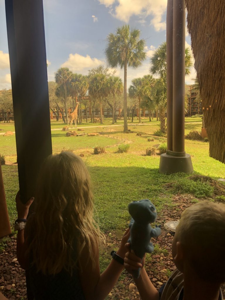 two children peer out a window at a savanna as a giraffe browses for leaves