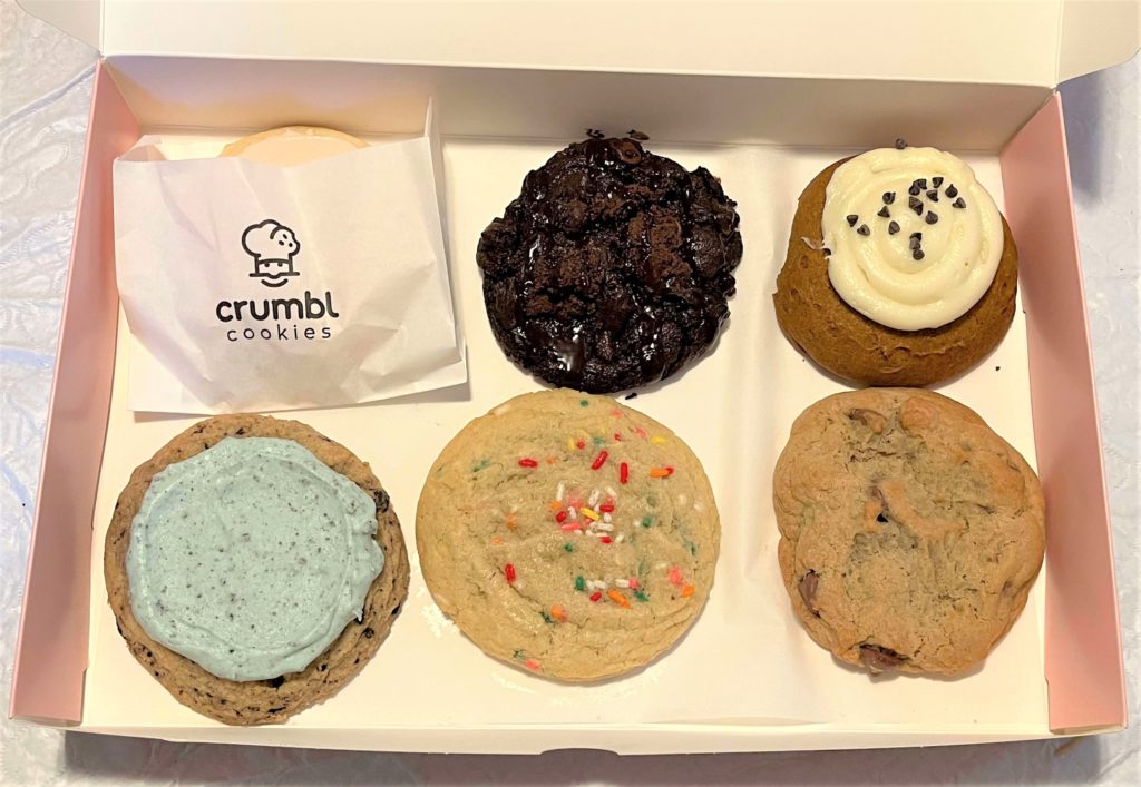 a variety pack of six jumbo cookies in an opened box bearing a smiling chef logo and the name Crumbl Cookies