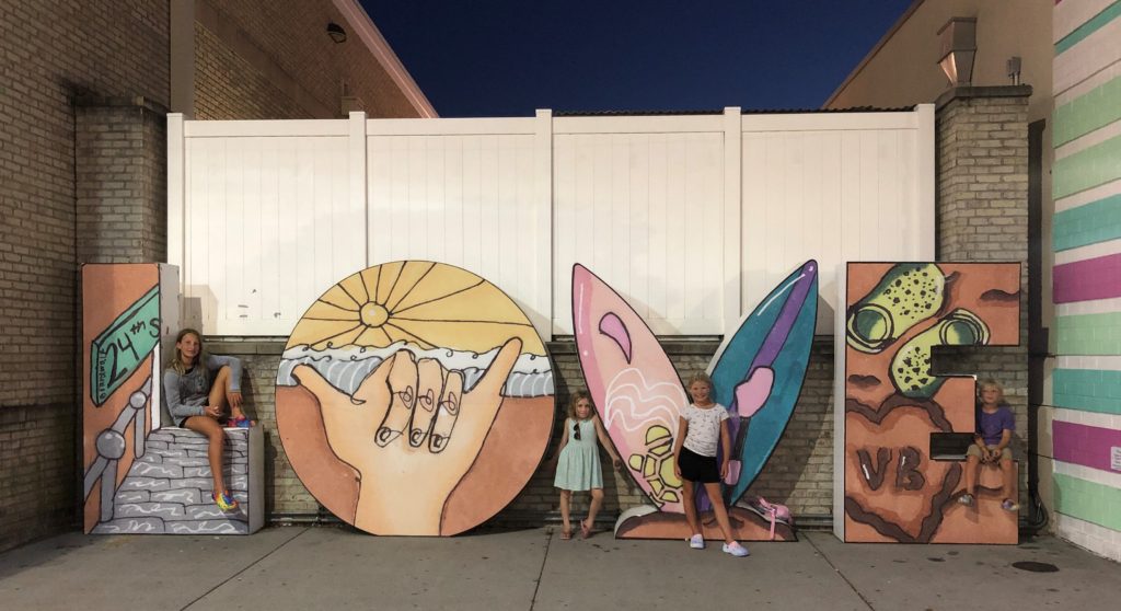 a giant lovework spells out L-O-V-E with 24th Street stairs, a "hang ten" sign, surfboards, and Crocs in the sand at the Virginia Beach oceanfront