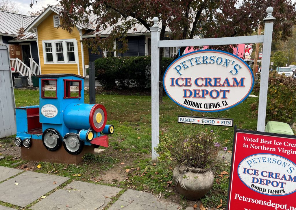 a tiny train engine sits outside of a large sign that reads "Peterson's Ice Cream Depot, Historic Clifton Virginia"
